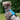 A curly-haired dog wearing a bright blue dog harness with black straps, sitting outdoors on a grassy lawn with green foliage in the background.