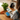A person holds a package of Fournblade Ocean Fish wet dog food over a wooden table, with a black cocker spaniel dog sitting on a chair in the background and fresh carrots and a green herb plant on the table.