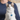 A white and brown cat wearing a blue collar with a KRUUSE silver tag that reads 'calm down', sitting on a person's lap. The person is wearing a navy blue long-sleeve shirt and is softly blurred in the background.
