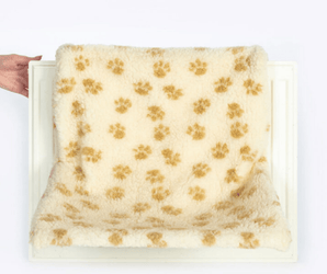 White background with a white radiator shown with a hand touch the side of it, on the radiator is a cream radiator cat bed with brown pawprints.