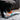 Black Labrador puppy with fox face dog toy made with soft orange fleece with a white fabric chin, smiley face stitch details; blue rope handles on each side in it's mouth stood on a dog bed with a brown sofa in the background.