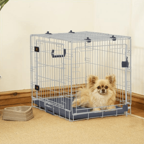 Cream coloured Pomeranian dog lying in a Rosewood Options dog crate in a corner of a room with a bowl next to it.