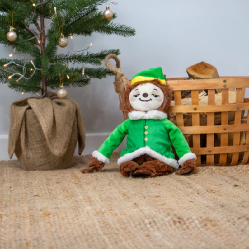 Room with a Christmas tree and a log basket with a brown sloth plush dog toy sat in front of it, wearing a green festive outfit and hat with stitched eye details and a cream face. 