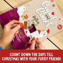 Hand opening a Rosewood Cupid and Comet Luxury Deli Dog Advent Calendar window with a dog treat in a wrapper. The advent calendar is beige and burgundy with two illustrated dogs pictured. At the bottom of picture is a red banner with white text 'Count down the days till Christmas with your furry friend!'