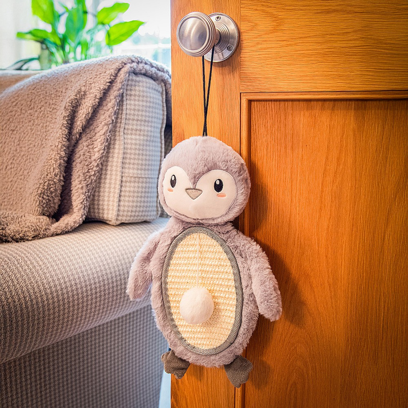 Penguin shaped cat scratcher, dominantly fluffy grey body with a canvas belly, with dark grey beak and feet, with a white pom pom on a string attached pictured hanging on a door knob next to a grey sofa. 