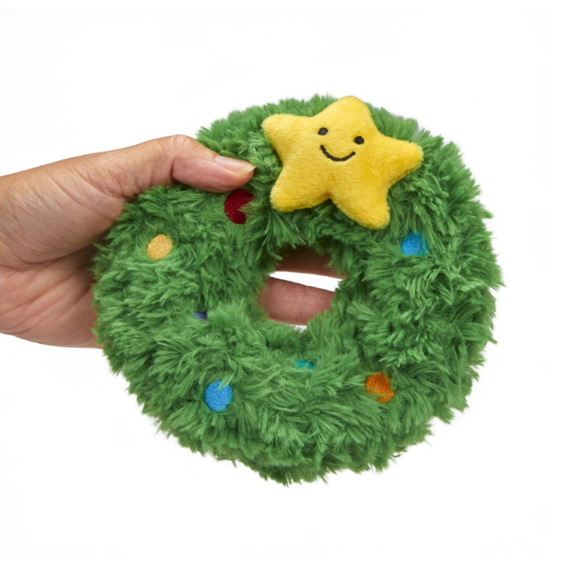White background with a hand holding a plush dog toy Christmas wreath with a plush star with stitched eyes and smile. 