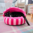 Black and white adult cat curled up inside a Sherpa pink stripy cat cave pictured in a living room. 
