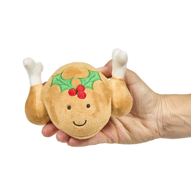 White background with a hand holding a plush dog toy roast turkey with a holly stitched on the top and stitched eyes and smile.