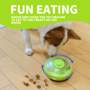 Brown and white collie-type dog playing with the UFO shaped treat dispenser with green central and top parts with main body in grey on a laminated floor. A bright green banner with white text say 'Fun Eating. Shove and chase the toy around to get the treats on the inside' 