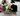 A black and white cat playing with an Ancol plush cat-shaped toy dressed as a bandit with a black-and-white striped shirt and a white bank box, on a living room carpet.
