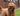 A brown poodle cross dog holding an Ancol Heritage plush bat-shaped dog toy made from brown fabric, in it's mouth. Pictured outside. 