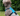 A curly-haired dog wearing a bright blue dog harness with black straps, sitting outdoors on a grassy lawn with green foliage in the background.
