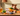 A senior collie dog sits behind a rustic wooden table displaying a box of Forthglade Turkey & Vegetable senior dog food, surrounded by fresh ingredients like carrots, pumpkin, parsley, and a blue bowl, in a warm, cozy room setting.