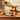 A senior collie dog sits behind a rustic wooden table displaying a box of Forthglade Turkey & Vegetable senior dog food, surrounded by fresh ingredients like carrots, pumpkin, parsley, and a blue bowl, in a warm, cozy room setting.