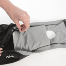 White background with one black bodysuit for dogs pictured with the back section open, showing the hole for the dog's tail, a hand is also pictured opening a pocket showing a pad inside. 