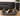 Livingroom scene with  a brown and white spaniel dog wearing a black Elizabethan style foam recovery collar with a grey rubber trim, lying in a grey dog bed. 