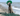 Beach scene with the ocean visible in the background, a Weimaraner in the foreground wearing a green Elizabethan style recovery collar with a green fishing net at its feet. 