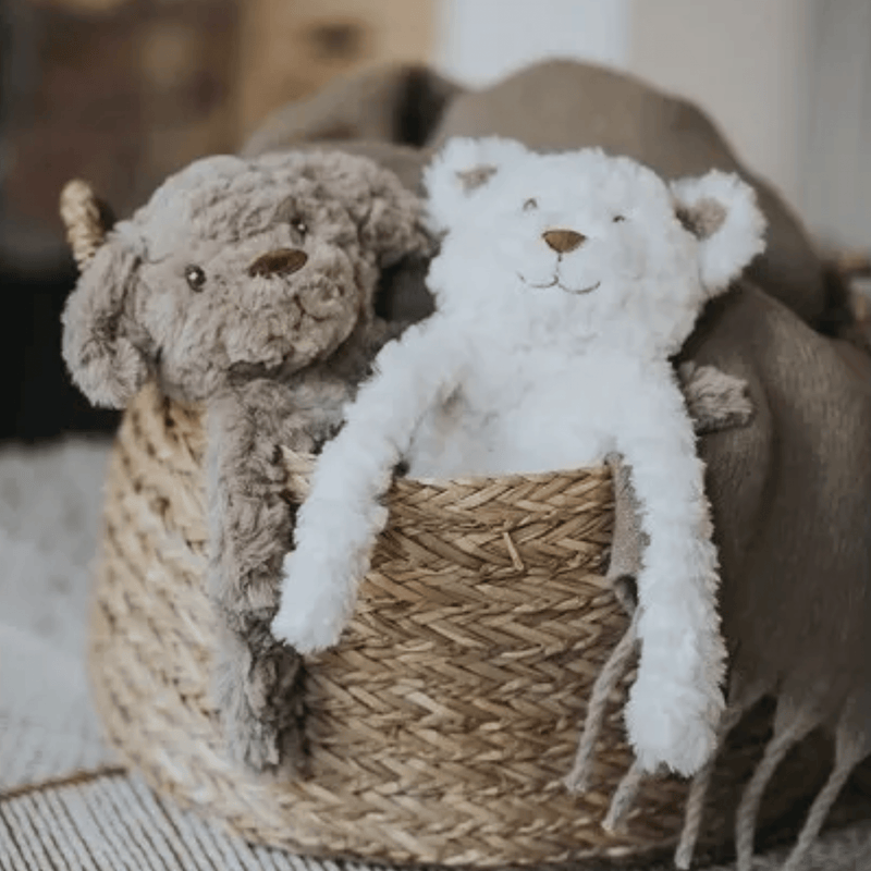 Close up of white bear shaped dog toy with long arms and legs, stitched detail eyes, brown nose pictured in a basket with the brown dog version of the same toy. 
