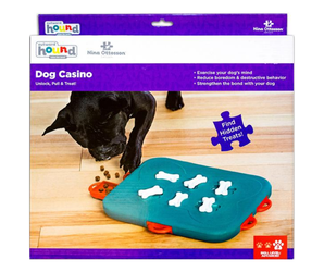 Nina Ottosson Hound Dog Casino puzzle toy in its retail packaging, featuring a teal board with white bone-shaped treats and red handles, shown on a white background.