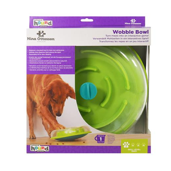 Nina Ottosson Wobble Bowl dog toy in its original retail packaging. The product is a bright lime green wobbly plastic bowl with a blue center button, shown through a clear window in a purple and white box featuring a picture of a golden retriever.