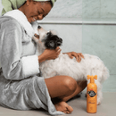 Black and white dog being held by a woman wearing a dressing grown using an orange bottle of Pet Head Ditch the Dirt shampoo in a tiled bathroom. 