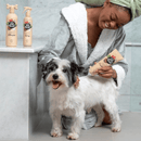 Black and white dog in front of an woman wearing a dressing grown using a a beige bottle of Pet Head Sensitive Soul conditioner in a tiled bathroom. On the shelf is a bottle of the shampoo and spray. 