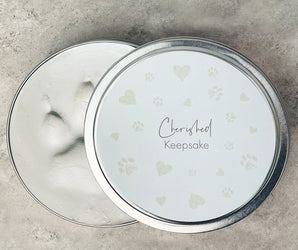 A round metal keepsake tin shown open, with the lid resting beside it. The lid is white with pale heart and paw-print motifs and the words 'Cherished Keepsake' in script and serif text.