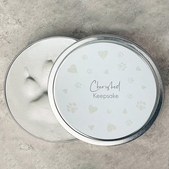A round metal keepsake tin shown open, with the lid resting beside it. The lid is white with pale heart and paw-print motifs and the words 'Cherished Keepsake' in script and serif text.