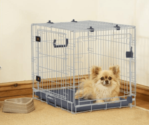 Cream coloured Pomeranian dog lying in a Rosewood Options dog crate in a corner of a room with a bowl next to it.
