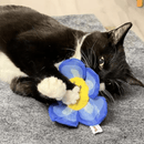 Black and white cat playing with blue flower cat toy with a yellow centre, with red stitched text that says 'Yeowww! catnip' on a grey rug. 